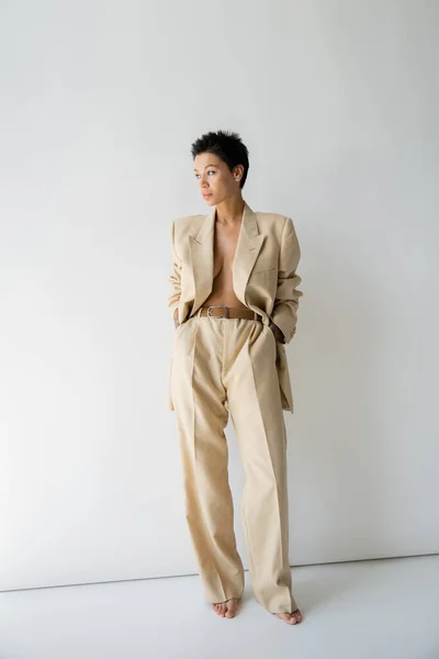 Full length of sexy barefoot woman in beige oversize blazer standing with hands in pockets and looking away on grey background — Stock Photo