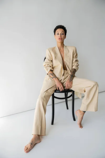 Full length of barefoot and sexy woman in oversize suit posing on chair and looking at camera on grey background — Stockfoto