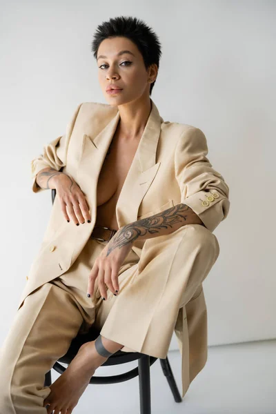 Séduisante femme tatouée en costume à la mode regardant la caméra tout en posant sur une chaise sur fond gris — Photo de stock