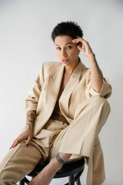 Sensual brunette woman in stylish suit holding hand near head while sitting on chair isolated on grey — Stock Photo