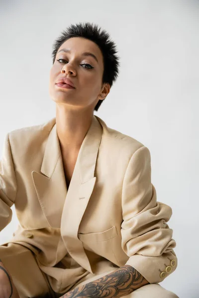 Portrait of brunette woman with makeup and piercing sitting in beige suit and looking at camera isolated on grey — Foto stock