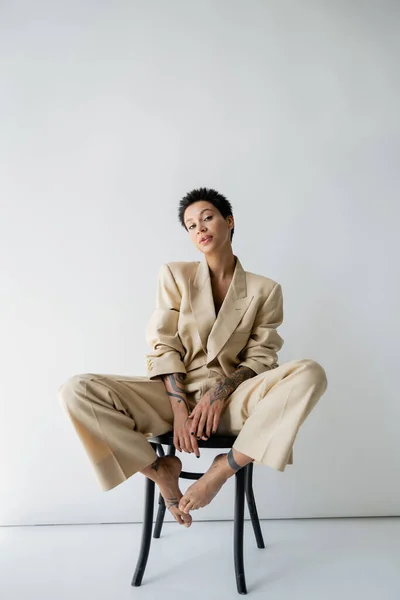 Full length of tattooed barefoot woman in stylish pantsuit posing on chair while looking at camera on grey background — Photo de stock