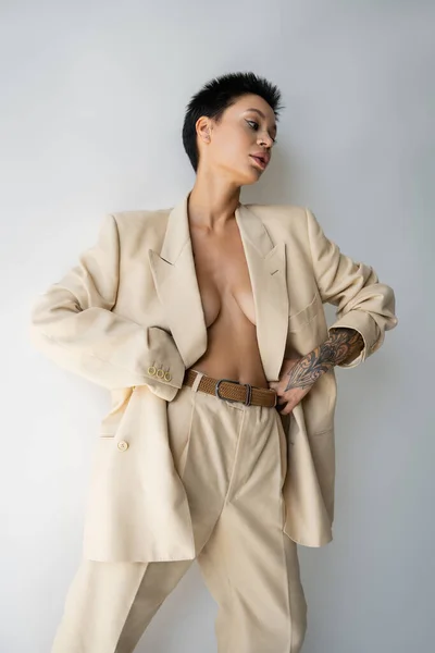 Tattooed woman wearing elegant oversize suit on sexy and shirtless body while standing with hands on waist on grey background — Stock Photo