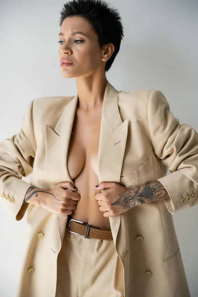 Sexy tattooed woman in beige blazer on shirtless body looking away on grey background — Stock Photo
