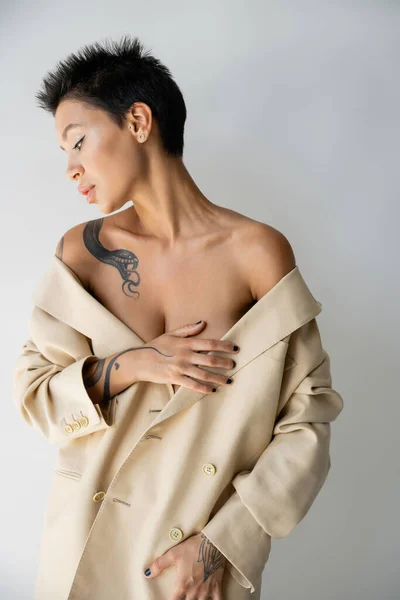 Sensual tattooed woman with bare shoulders covering breast with hand while posing in oversize blazer isolated on grey — Stock Photo