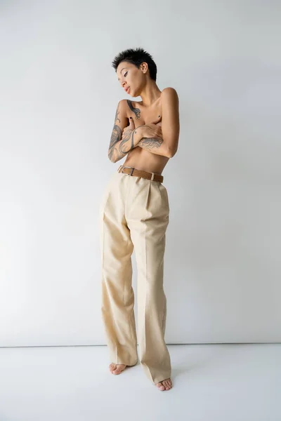 Full length of sexy shirtless woman in beige pants hiding breast with hands on grey background — Stock Photo
