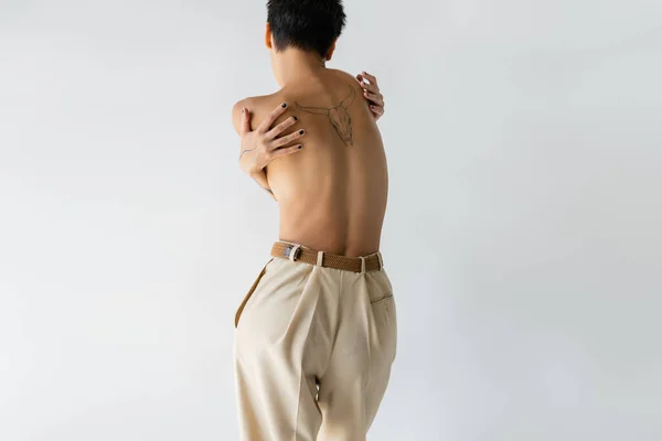 Back view of tattooed shirtless woman in beige pants hugging herself isolated on grey — Stock Photo