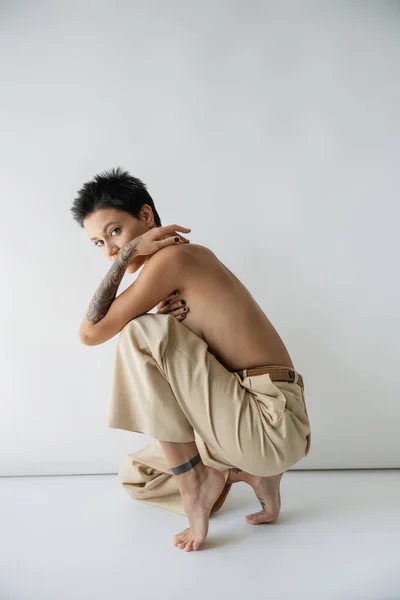 Shirtless barefoot woman in beige pants looking at camera on grey background — Foto stock