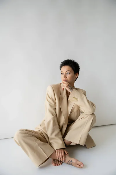Pensive barefoot woman in beige suit sitting with crossed legs and looking away on grey background — Stock Photo