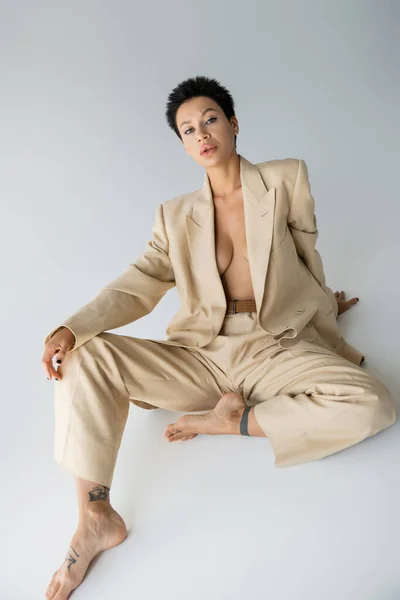 Shirtless and barefoot woman in elegant pantsuit looking at camera while sitting on grey background — Stock Photo