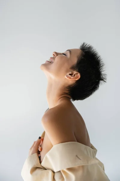 Vista laterale di donna felice e sexy in blazer beige con spalle nude che coprono il seno con le mani isolate sul grigio — Foto stock