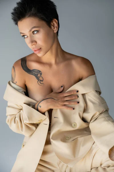 Sexy woman with tattoo on shirtless body posing with beige blazer isolated on grey — Fotografia de Stock