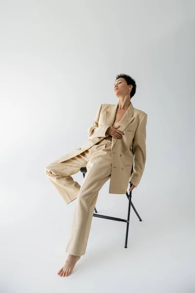 Full length of barefoot woman wearing elegant suit on shirtless body and posing on chair on grey background — Stock Photo