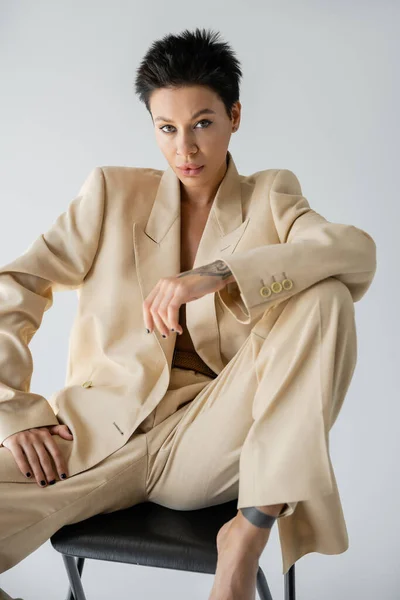 Tattooed woman in elegant oversize suit posing on chair and looking at camera isolated on grey — Stock Photo