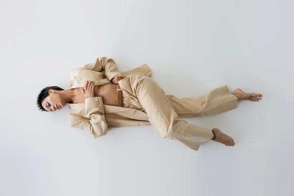 Vista dall'alto della donna scalza e senza maglietta in abito beige che copre il seno con la mano mentre giace su sfondo grigio — Foto stock