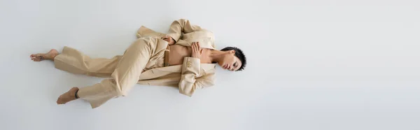 Top view of barefoot and shirtless woman in beige pantsuit lying on grey background, banner — Stock Photo