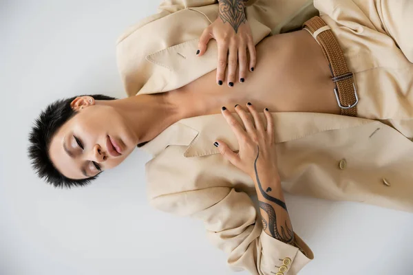 Top view of sexy woman in beige blazer covering breast with hands while lying with closed eyes on grey background — Foto stock