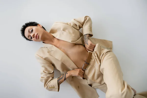Top view of seductive tattooed woman in pantsuit on shirtless body lying on grey background — Stock Photo