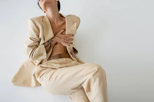 Cropped view of sexy shirtless woman covering breast with hand while sitting in elegant pantsuit on grey background — Stock Photo