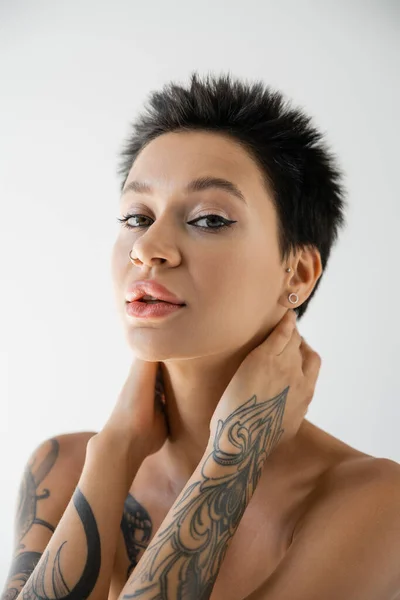 Ritratto di donna tatuata con trucco e spalle nude che tiene le mani sul collo e guarda la fotocamera isolata su grigio — Foto stock
