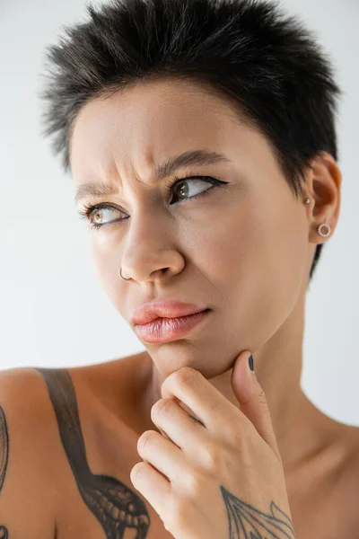 Portrait of upset and frowning woman with makeup and piercing touching chin and looking away isolated on grey — Stock Photo