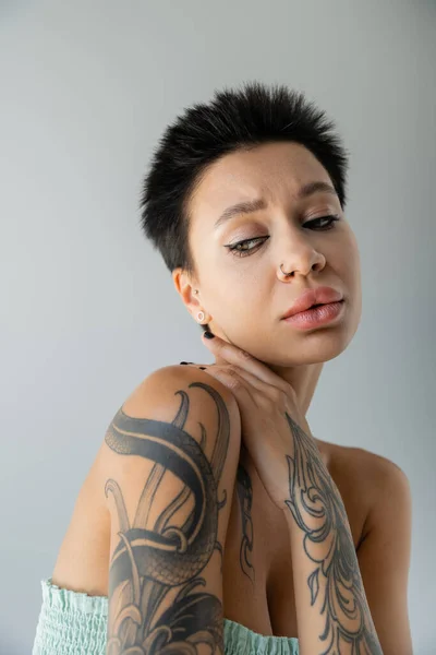 Portrait of upset tattooed woman in strapless top posing isolated on grey — Stock Photo