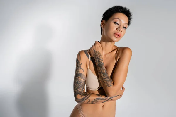 Tattooed and sensual woman in bra looking at camera while posing on grey background — Fotografia de Stock