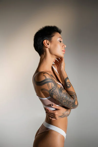 Side view of tattooed woman with short brunette hair touching face while standing in bra on grey background — Fotografia de Stock