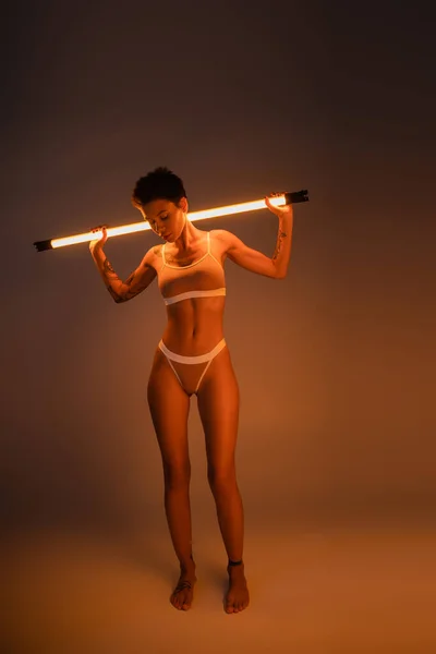 Comprimento total de mulher tatuada descalça em roupa interior em pé com lâmpada vibrante no fundo escuro — Fotografia de Stock