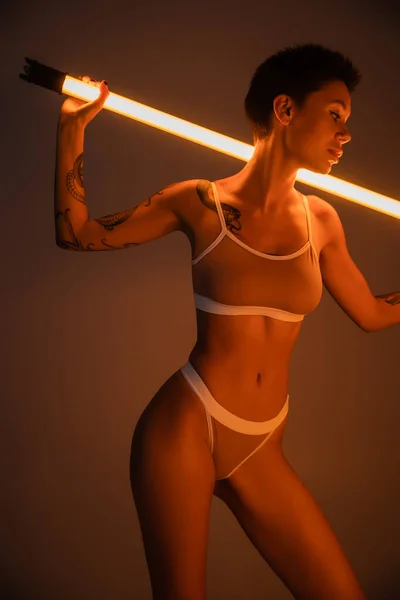 Verführerische Frau mit perfekt tätowiertem Körper posiert in Unterwäsche mit Leuchtlampe auf dunklem Hintergrund — Stockfoto