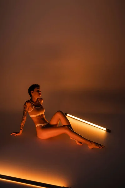 Full length of slender tattooed woman in lingerie sitting near vibrant fluorescent lamps on dark background — Photo de stock