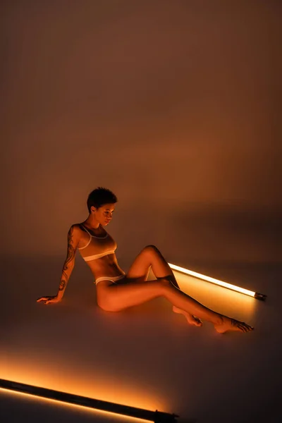 Full length of tattooed woman in underwear sitting near fluorescent lamps on dark background — Photo de stock