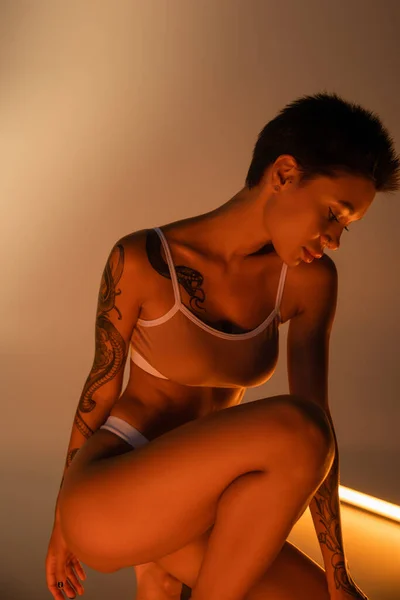 Сексуальна татуйована жінка в нижній білизні позує у світлі люмінесцентної лампи на бежевому фоні — стокове фото