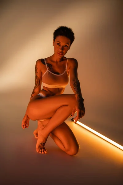 Полная длина молодая женщина с идеальным телом, смотрящая на камеру возле флюоресцентной лампы на бежевом фоне — стоковое фото