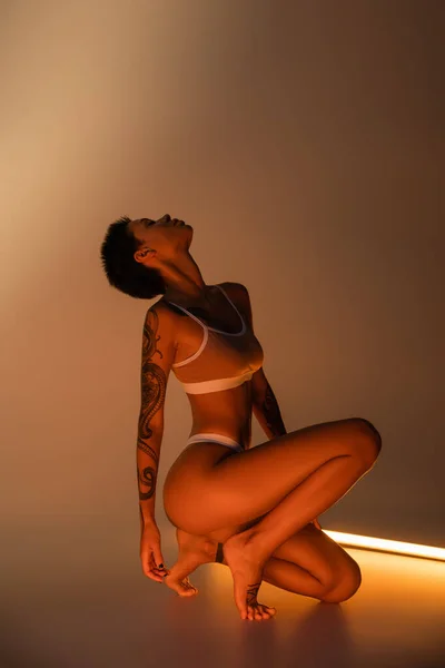 Side view of fit woman with perfect tattooed body posing in underwear near fluorescent lamp on beige background — Stock Photo
