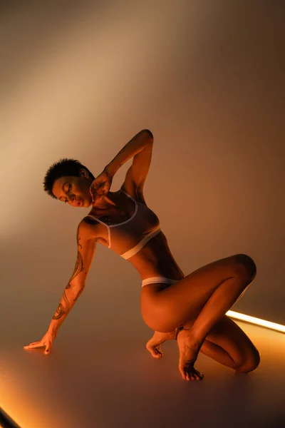 Volle Länge der verführerischen Frau mit tätowierten und perfekten Körper posiert in Unterwäsche in der Nähe von Leuchtstoffröhren auf beigem Hintergrund — Stockfoto