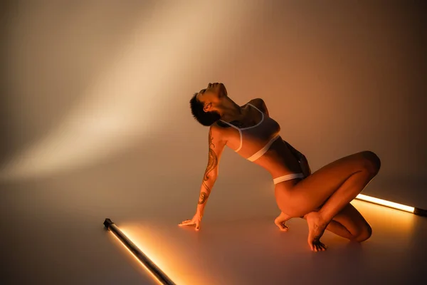 Comprimento total de mulher tatuada morena em roupa interior posando perto de lâmpadas fluorescentes no fundo bege — Fotografia de Stock