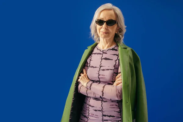 Senior woman in stylish outfit and sunglasses standing with crossed arms isolated on blue — Stock Photo