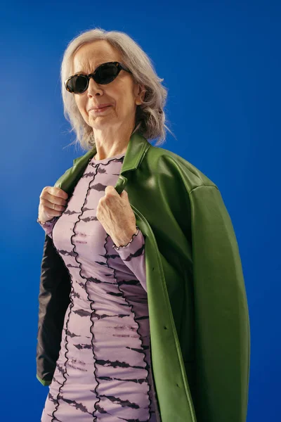 Satisfied senior woman in sunglasses wearing green leather jacket isolated on blue — Stock Photo
