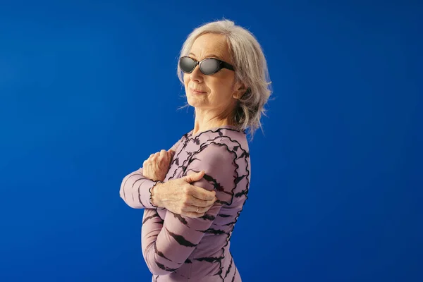Cheerful senior woman in sunglasses and dress looking at camera isolated on blue — Stock Photo