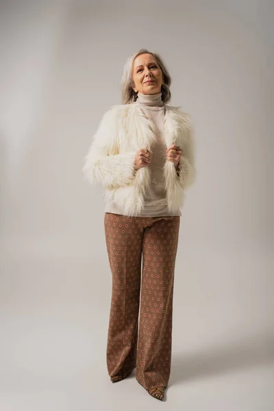 Full length of pleased senior woman in faux fur jacket posing on grey background — Stock Photo