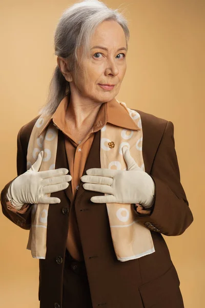 Senior woman in brown formal wear and white gloves posing isolated on beige — Stock Photo