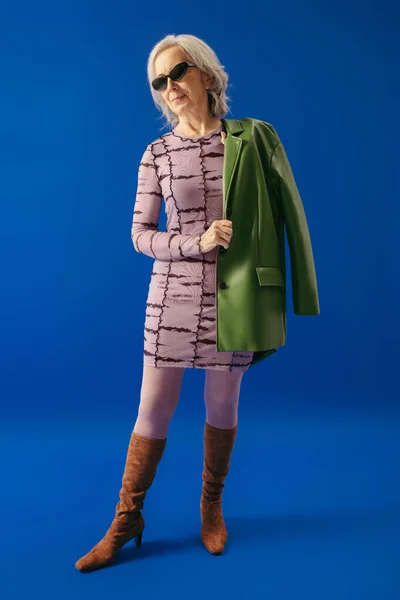 Full length of senior woman in dress and boots standing with green jacket on blue — Foto stock