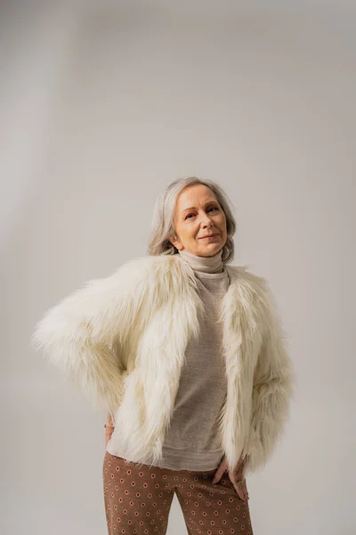 Pleased elderly woman in white faux fur jacket posing with hand on hip on grey — Photo de stock