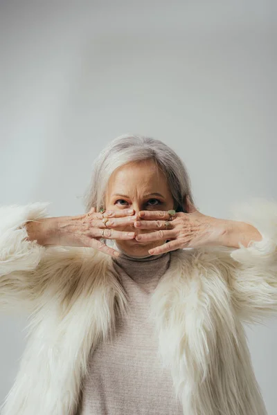 Portrait of senior woman with rings on fingers looking at camera while covering face with hands on grey — Stock Photo