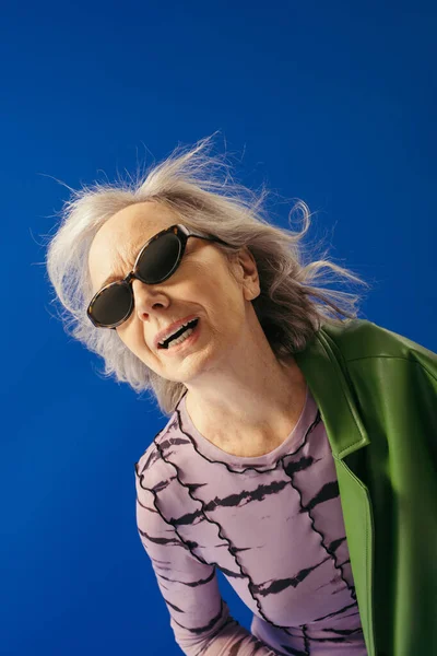 Senior woman in trendy sunglasses and green leather jacket standing against wind isolated on blue — Stock Photo