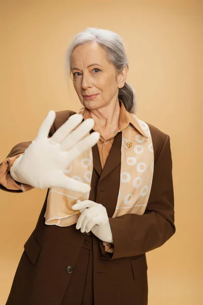 Elderly woman in white gloves looking at camera while showing stop sign isolated on beige — Stock Photo