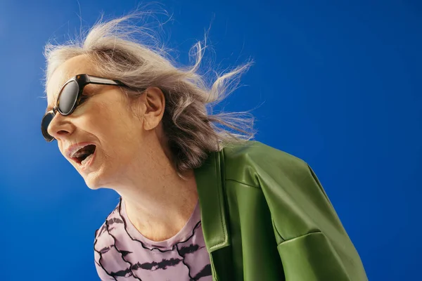 Senior woman in trendy sunglasses and green leather jacket standing with opened mouth against wind isolated on blue — Stock Photo
