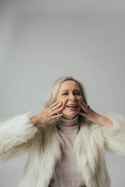 Portrait de femme âgée joyeuse avec des anneaux sur les doigts regardant la caméra tout en touchant le visage sur le gris — Photo de stock