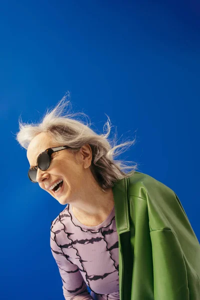 Happy senior woman in trendy sunglasses and green leather jacket laughing against wind isolated on blue — Stock Photo